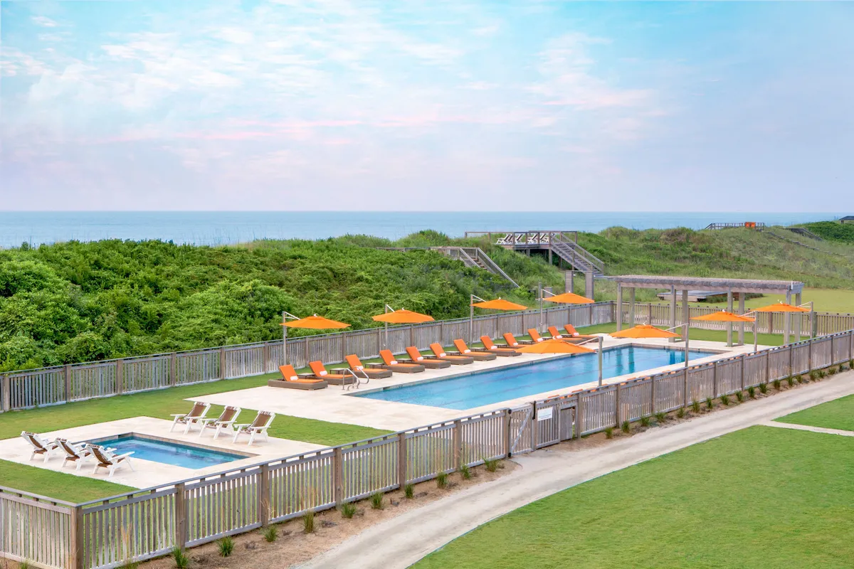 A beautifully landscaped outdoor pool area features inviting lounge chairs, vibrant orange umbrellas, and lush greenery. A clear pathway leads directly to the beach, merging the aquatic amenities with seaside relaxation. The space is alive with families and friends relaxing, sunbathing, and playing outdoor games, encapsulating the lively, communal spirit of the resort.