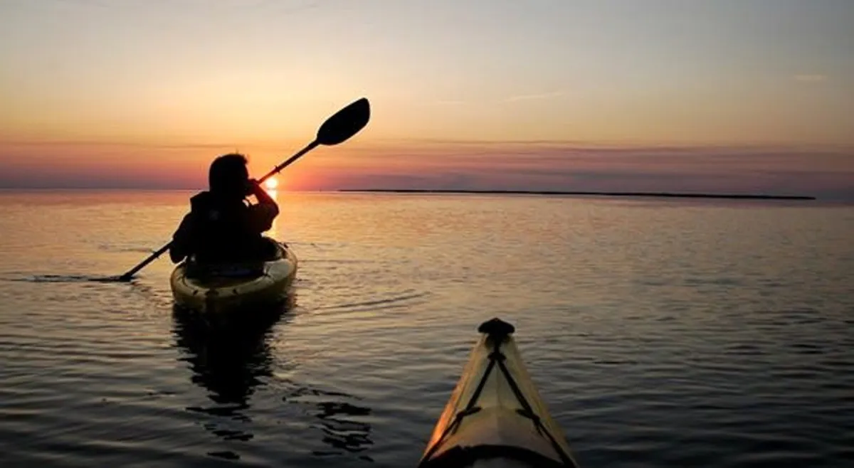 A kayaker glides across a calm sound at sunset, with a vivid reflection of the sky's colors on the water's surface. The tranquil, expansive view conveys a sense of adventure and quiet intimacy set against a backdrop of natural beauty, invoking the idea of exploring Currituck Sound together.