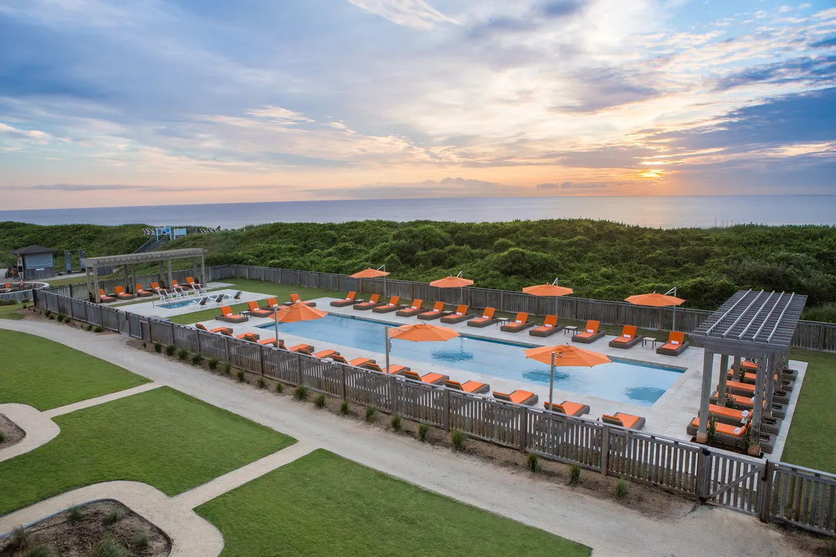 An inviting swimming pool area at sunset, with lounge chairs and orange umbrellas lining the water's edge. The tranquil water reflects the sky's golden hues, suggesting a peaceful yet vibrant atmosphere ideal for unwinding. The scene exudes relaxation and is perfect for couples to enjoy a romantic evening or celebrate together.
