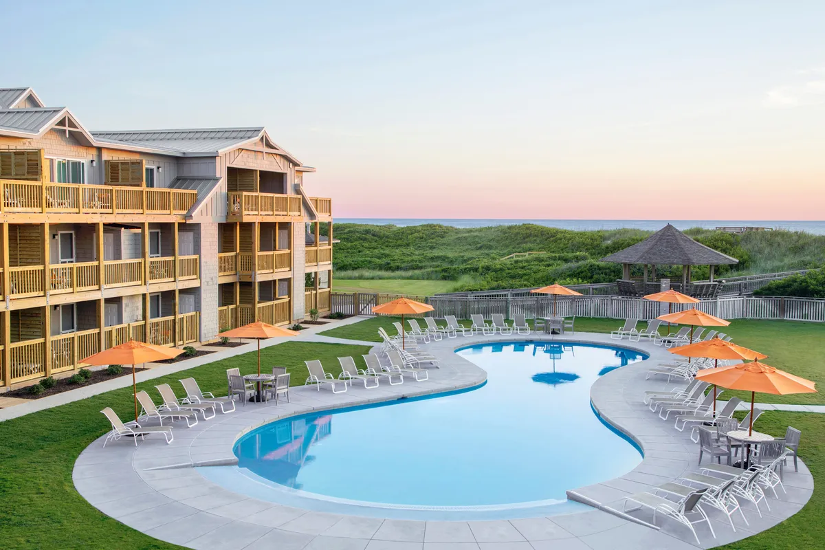 A vibrant outdoor pool area with sparkling blue water, orange umbrellas, and a well-kept lawn. Guests are seen swimming, sunbathing, and relaxing around the pool, with the resort facade providing an inviting backdrop.
