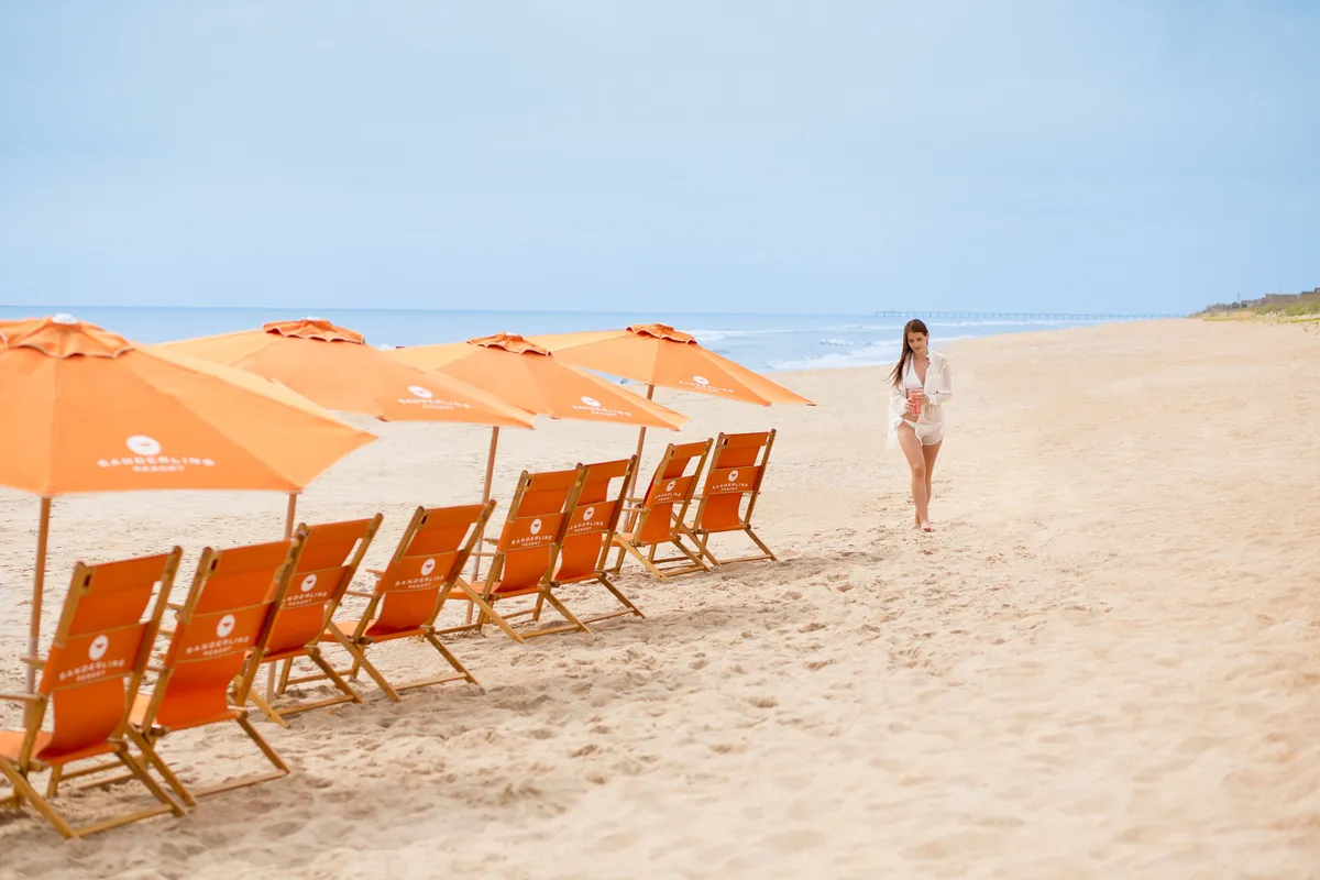 This image showcases a tranquil beach scene with beach chairs and umbrellas set along the shore, the ocean stretching into the distance, and a woman walking nearby. The open sandy space is inviting and evokes the freedom and relaxation of a seaside escape, perfect for morning walks or unwinding after a day of adventure.