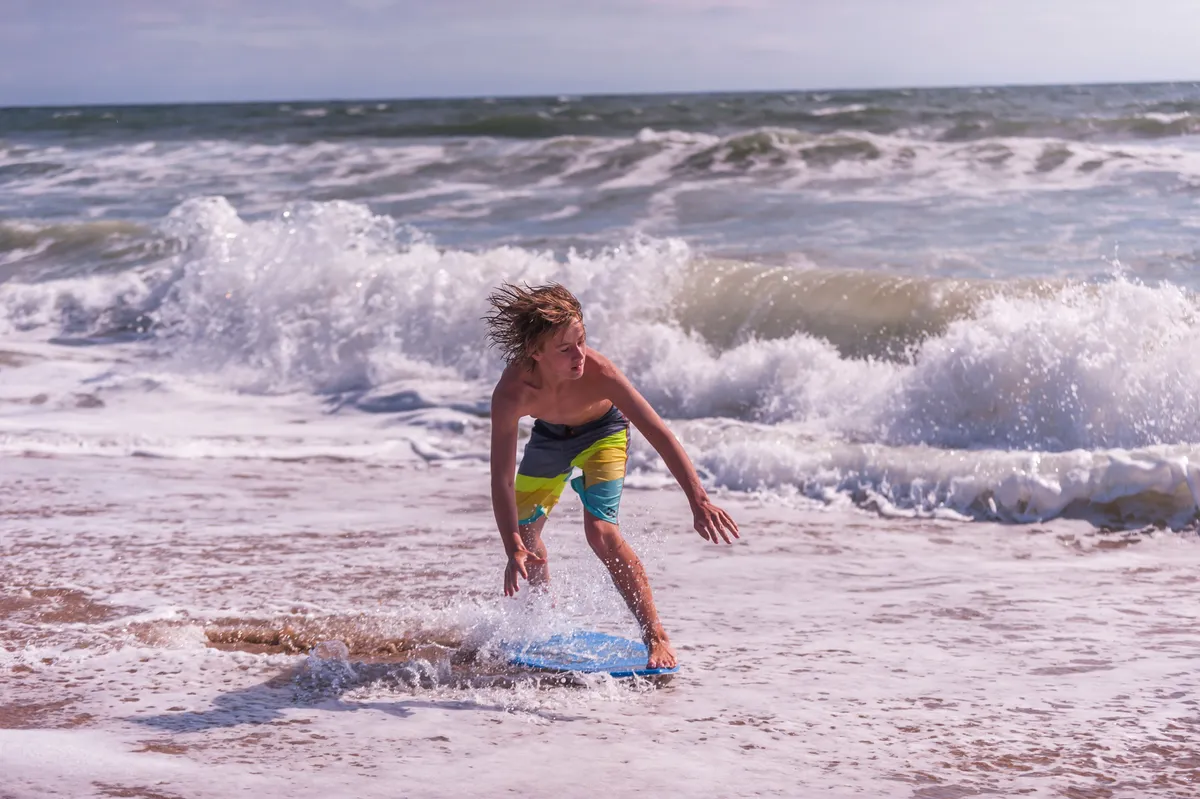 A young boy gleefully rides a boogie board on gentle waves, with sunlight sparkling across the water. The shoreline and ocean environment evoke an energetic and playful atmosphere, emblematic of family-friendly beach adventures.