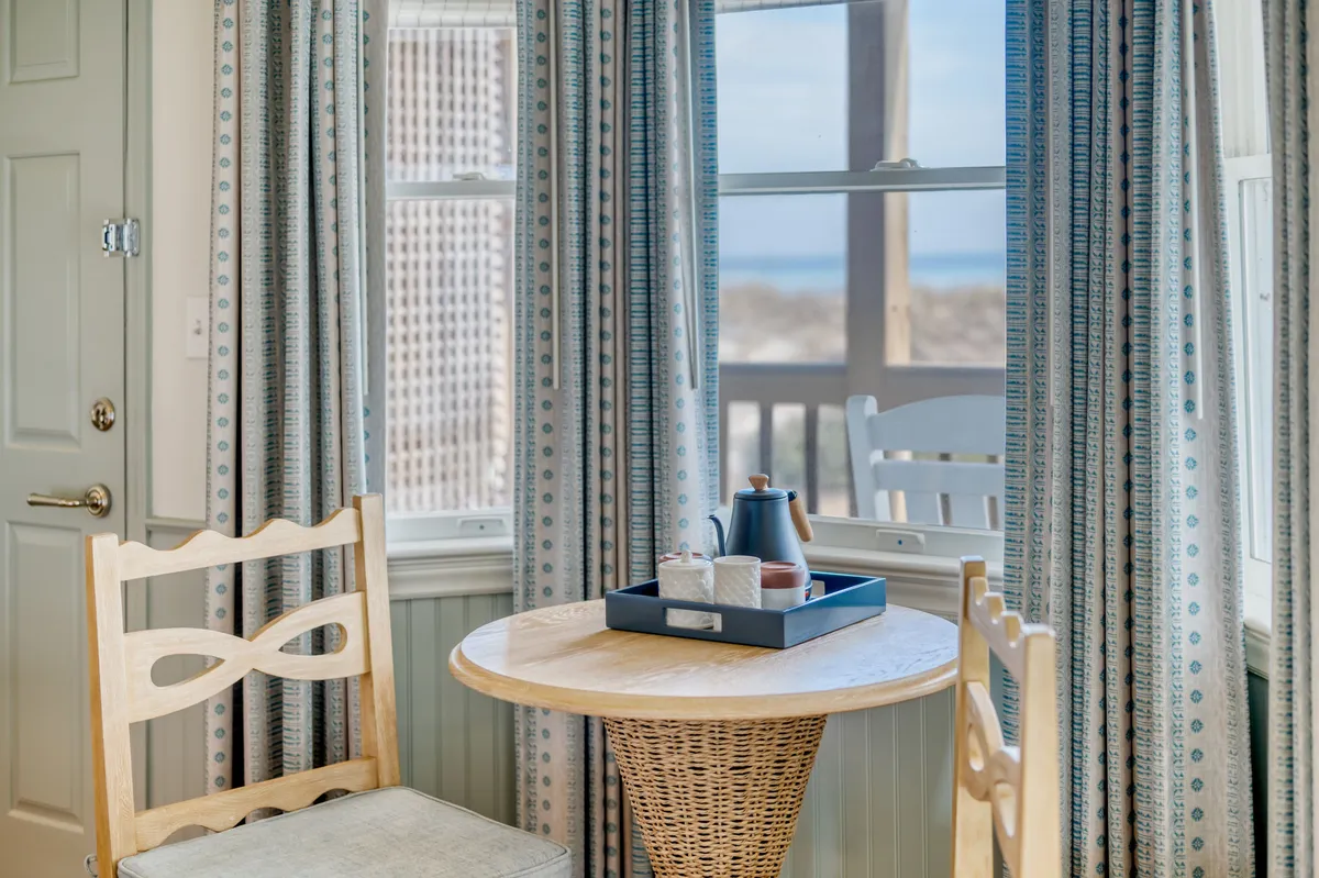 A cozy sitting nook featuring a wooden chair, a small round table set with a tea set, curtains, and a window offering a peaceful view. The scene suggests an inviting spot for morning coffee, relaxation, or intimate board games.