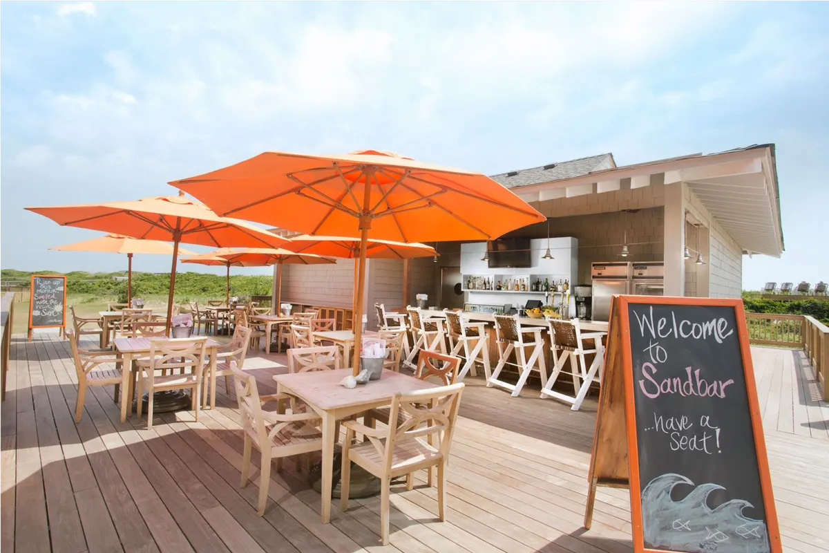 An outdoor bar and dining area with cheerful orange umbrellas and wooden tables set just beside the beach. The atmosphere is lively and inviting, with the ocean visible in the background, perfect for enjoying cocktails and each other's company as the sun sets.