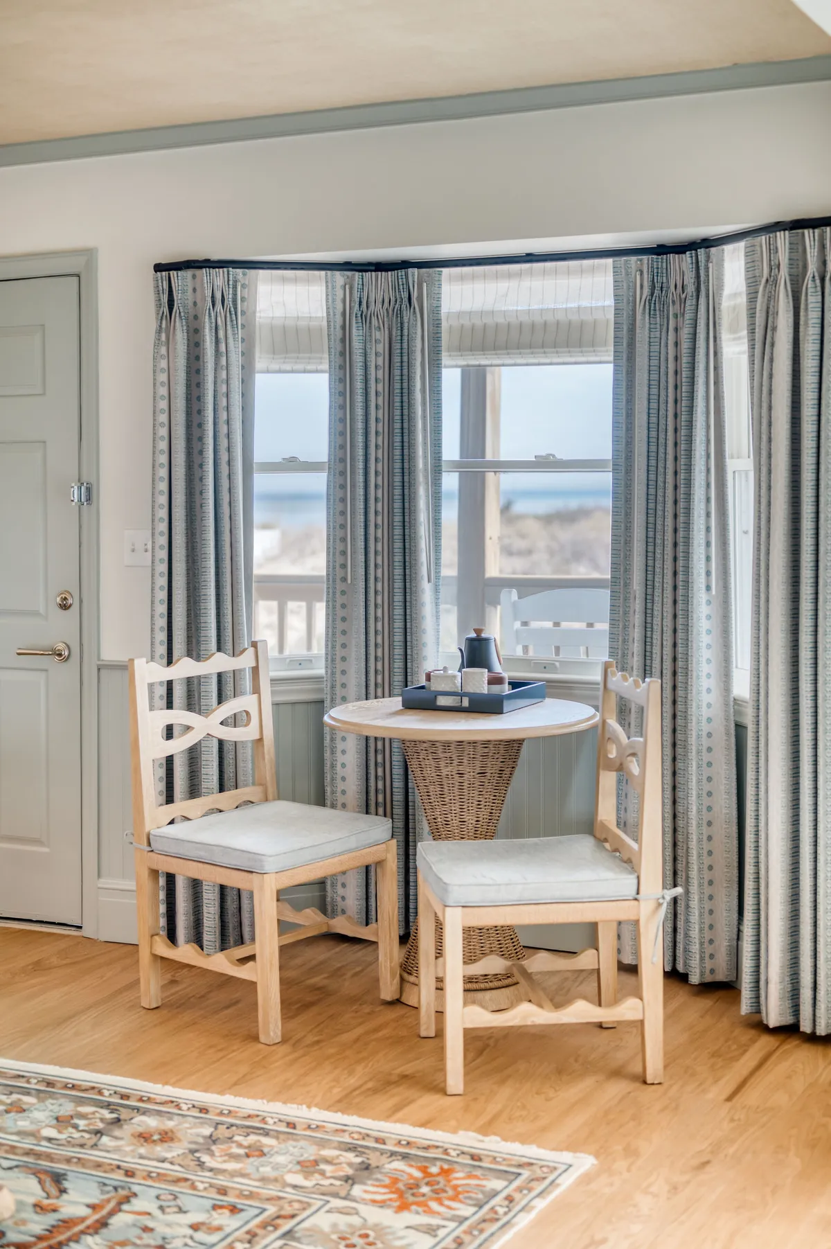 A cozy interior setting with a round table and two wooden chairs positioned beside a window, sheer curtains filtering soft daylight. Outside the window, you can glimpse an inviting view, suggestive of an oceanfront perspective. The scene exudes tranquility and intimacy—perfect for reading or enjoying a morning coffee while taking in the sunrise.