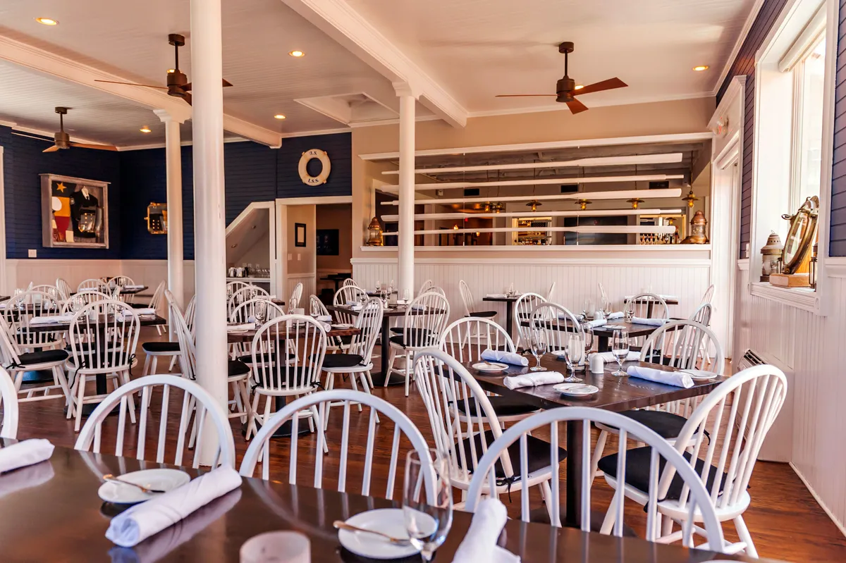 A bright, inviting dining area featuring wooden tables, white chairs, a stylish bar area, and decorative wall art. The space exudes a welcoming, modern atmosphere where guests can gather to enjoy meals and socialize. Natural light highlights the clean lines and coastal-inspired decor, connecting the interior ambiance with the relaxed Outer Banks environment.
