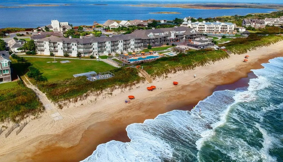A panoramic beachfront scene shows the sparkling swimming pool adjacent to the sandy shore, with colorful umbrellas and the resort's architecture creating a festive, holiday atmosphere. Families are gathered poolside, while others enjoy sunbathing, beach sports, and playful moments on the sand, emphasizing the full spectrum of resort activities available.