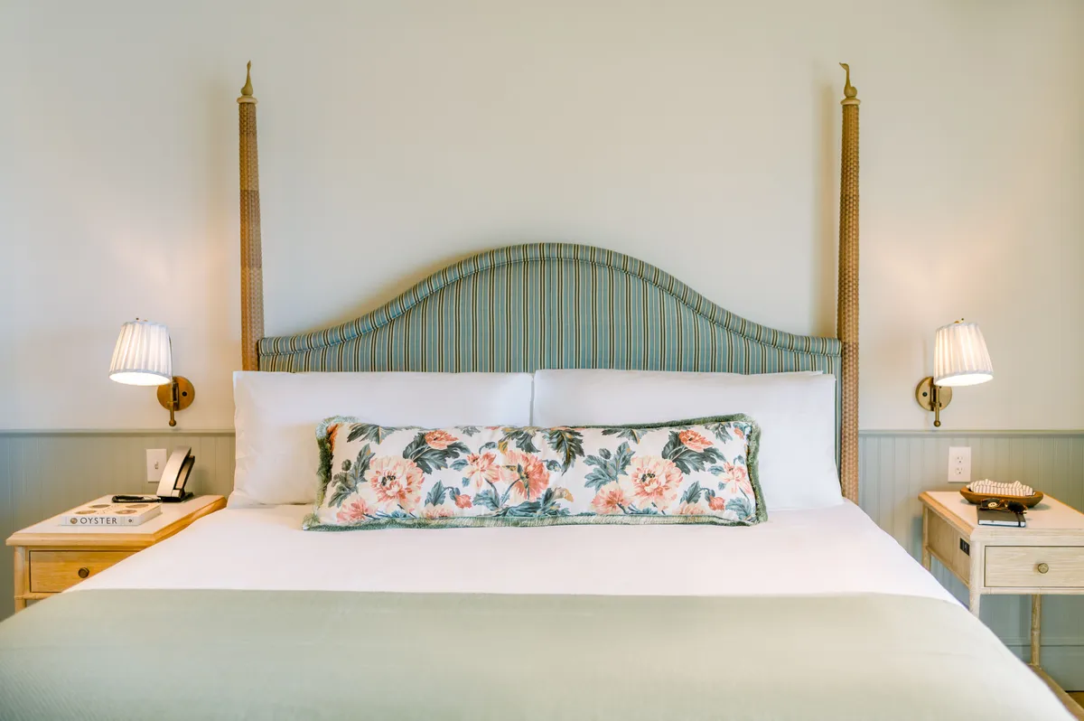 A tranquil bedroom space with a plush bed dressed in luxury linens, a floral pillow, wall-mounted lights, and a nightstand with decorative items. The overall aesthetic is cozy and inviting, perfectly suited for restful nights and morning relaxation.