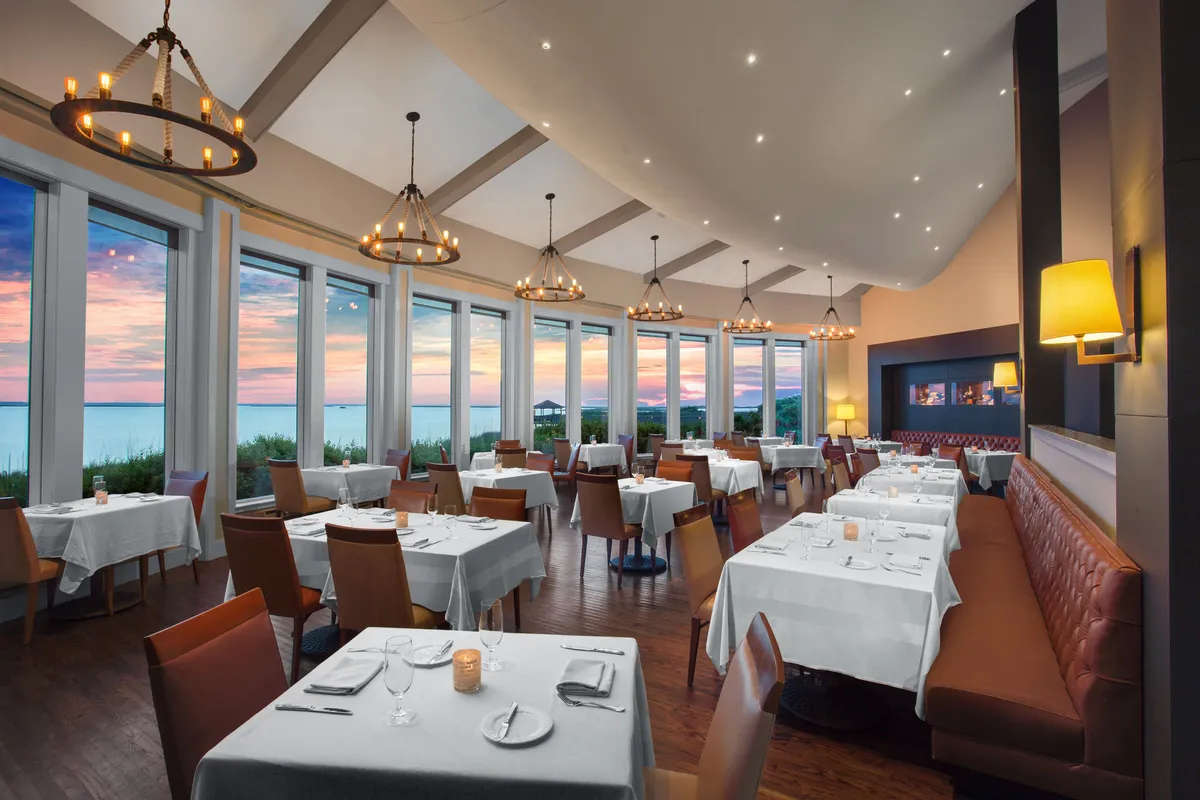 A sophisticated dining space with large windows frames tranquil sunset views over the water. Well-set tables and elegant lighting create an inviting atmosphere, perfect for romantic dinners or memorable family meals, capturing the essence of on-site dining with a view.
