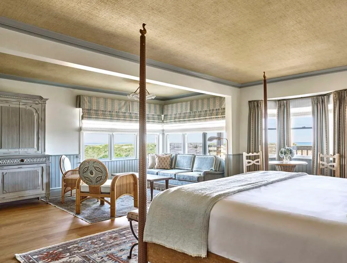 The image showcases a bright, elegant studio interior with a luxurious four-poster king bed dressed in fine linens, a cozy blue sofa seating area, a sleek wooden cabinet, and expansive windows letting in abundant natural light and offering panoramic Atlantic Ocean views.